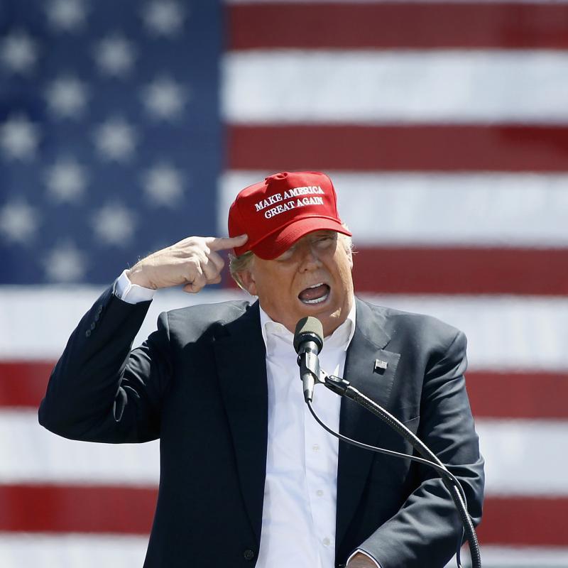 Donald Trump speaking at a campaign event in a MAGA hat with an American flag backdrop