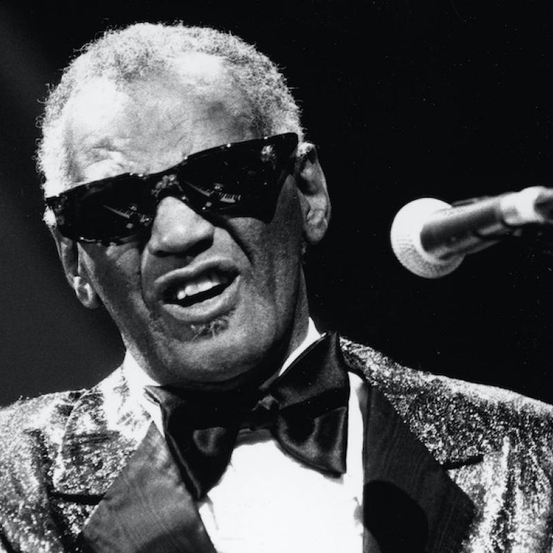 Legendary musician Ray Charles sits at his piano and sings into a mic while wearing his trademark sunglasses in this black and white image from 1985